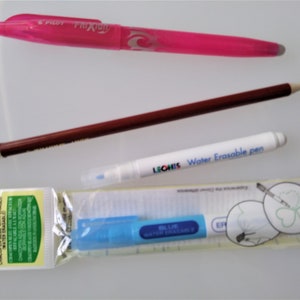 May include: A close-up of four different marking tools: a pink erasable pen, a brown pencil, a white water erasable pen, and a blue water erasable pen in a plastic package. The blue pen has the text "BLUE WATER ERASABLE" printed on it. The white pen has the text "LEONIS Water Erasable pen" printed on it. The package has the text "Clover Art No. 5013 CHACOPEN BLUE WITH ERASER" printed on it.