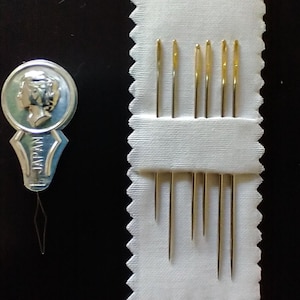 K&ouml;nnte beinhalten: Ein Set goldfarbener N&auml;hnadeln in einem wei&szlig;en Stoffhalter, zusammen mit einem Nadeleinf&auml;dler. Der Einf&auml;dler ist silber und blau, mit dem Aufdruck "JAPAN". Die Nadeln sind unterschiedlich lang.