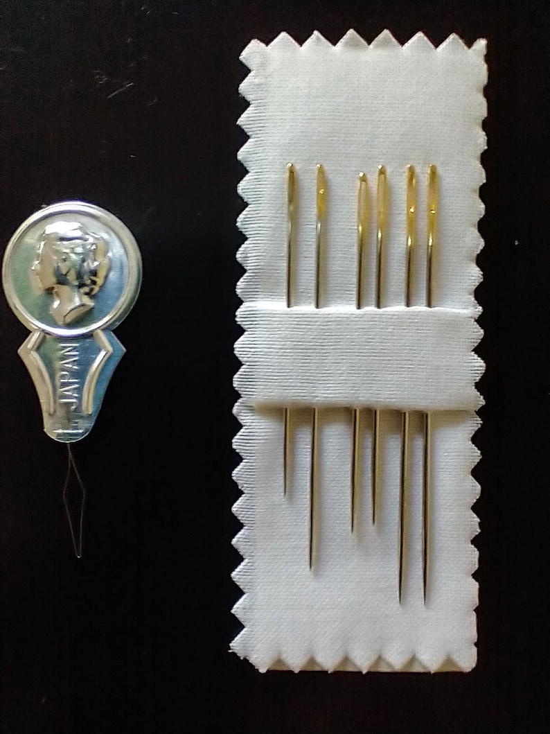 K&ouml;nnte beinhalten: Ein Set goldfarbener N&auml;hnadeln in einem wei&szlig;en Stoffhalter, zusammen mit einem Nadeleinf&auml;dler. Der Einf&auml;dler ist silber und blau, mit dem Aufdruck "JAPAN". Die Nadeln sind unterschiedlich lang.
