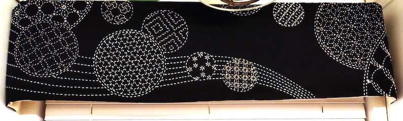 Puede incluir: Cenefa de tela negra con patrones circulares bordados en blanco y l&iacute;neas punteadas. El dise&ntilde;o presenta c&iacute;rculos de varios tama&ntilde;os con costuras detalladas, creando un aspecto decorativo y texturizado.