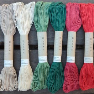 May include: Six skeins of Sashiko embroidery thread in beige, off-white, sage green, dark green, magenta, and red. Each skein is wrapped in a tan paper band featuring the text Sashiko, 20m, Cotton 100%, and Made in Japan in black ink.