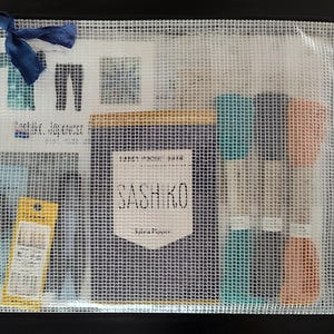 K&ouml;nnte beinhalten: Eine transparente Netztasche mit verschiedenen Bastelmaterialien. Darin befinden sich Garnrollen in T&uuml;rkis und Pfirsich, ein Nadelpaket und eine marineblaue Schleife. Die Tasche enth&auml;lt auch eine Karte mit dem Text "Sashiko, Japanese..." und ein Etikett mit der Aufschrift "SASHIKO".
