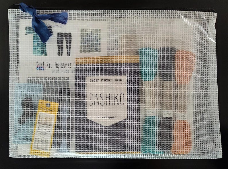 K&ouml;nnte beinhalten: Eine transparente Netztasche mit verschiedenen Bastelmaterialien. Darin befinden sich Garnrollen in T&uuml;rkis und Pfirsich, ein Nadelpaket und eine marineblaue Schleife. Die Tasche enth&auml;lt auch eine Karte mit dem Text "Sashiko, Japanese..." und ein Etikett mit der Aufschrift "SASHIKO".