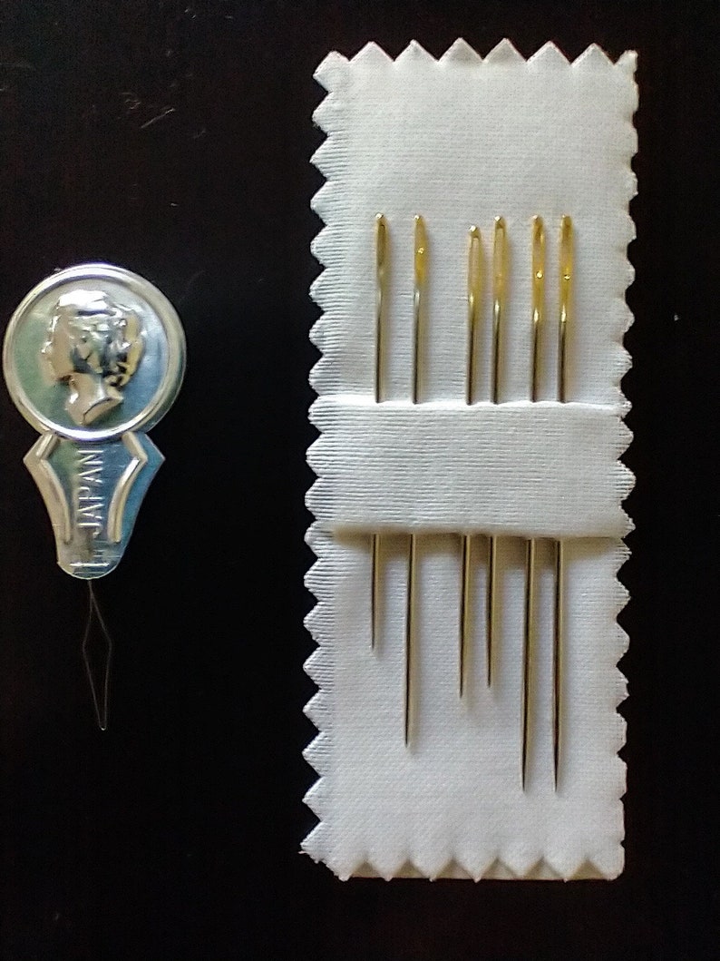 Op de afbeelding: Een set van acht goudkleurige naalden in een wit stoffen etui met gegolfde randen. Het etui heeft de tekst "JAPAN" erop gedrukt.