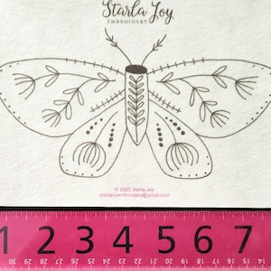 May include: A cream-colored fabric square featuring a detailed, brown line drawing of a butterfly with floral and dotted wing patterns. The text "Starla Joy EMBROIDERY" is at the top. A pink ruler with inch measurements is at the bottom.