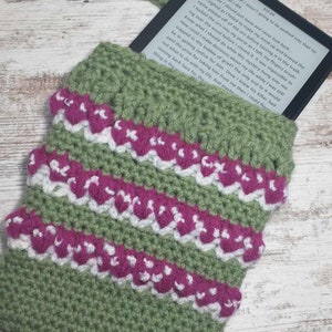 May include: Green crocheted e-reader case with pink and white accents. The case has a strap and is holding an e-reader.