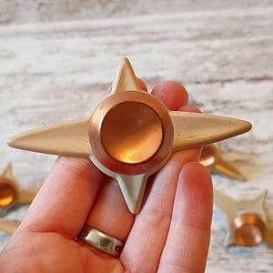 May include: Cream-colored star-shaped cabinet knobs with copper-colored center accents. The knobs have a four-point star design. The image shows several knobs, with one held in a hand. The background is a light-colored wooden surface.