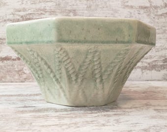 May include: A light green ceramic bowl with a geometric shape. The bowl has a textured, embossed design on the sides and a smooth, flat rim. The bowl is sitting on a white wooden surface.