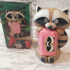 May include: A raccoon-shaped soap dispenser holding a pink bar of soap. The dispenser is brown, white, and black, with a cartoonish design. The soap bar has a raccoon graphic and the text "SOAP NET WT 3 OZ." A matching box is visible.