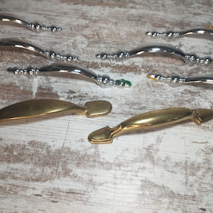 May include: A collection of cabinet handles in various styles. Some are shiny silver with decorative bead accents and others are a brushed gold color. The handles are curved and have a classic design, suitable for furniture restoration or new builds.