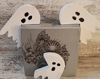 May include: Three white wooden ghost decorations with black dot eyes are arranged around a gray box with an ornate design. The ghosts have a simple, cartoonish design, perfect for Halloween or spooky decor.