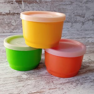 May include: Three colorful plastic containers with lids. The containers are stacked, with a yellow container on top, a green one in the middle, and an orange one at the bottom. Each container has a matching lid in a light peach color.