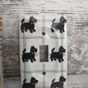 May include: White ceramic light switch plate with a plaid pattern and black silhouettes of Scottish Terriers wearing red bow ties.