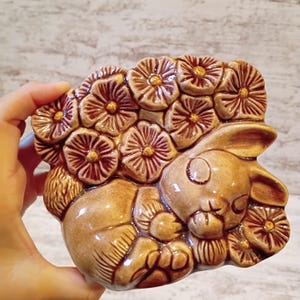 May include: A glazed ceramic figurine featuring a sleeping rabbit nestled among a cluster of sculpted flowers. The piece is finished in warm brown tones with darker accents on the petals and a glossy, reflective surface throughout the design.