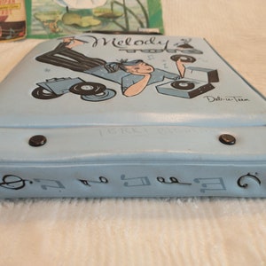 May include: A light blue vinyl record carrying case with a black and white illustration of a woman listening to records. The case has the words "Melody Time" and "Del-in-Teen" printed on it. The case has two black metal clasps.