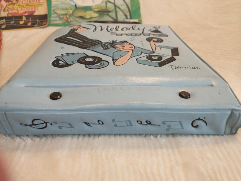May include: A light blue vinyl record carrying case with a black and white illustration of a woman listening to records. The case has the words "Melody Time" and "Del-in-Teen" printed on it. The case has two black metal clasps.