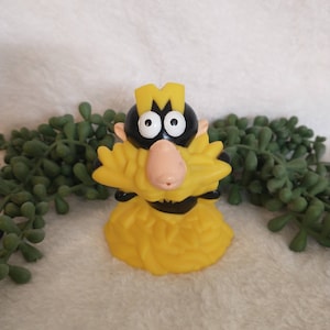 May include: A yellow and black plastic toy figure of a cartoon character with a large mustache and a letter "M" on its head.