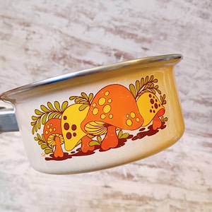 May include: Cream-colored enamel saucepan with a silver rim and handle. The side features a colorful illustration of orange and yellow mushrooms with green foliage. A charming, vintage kitchenware item.