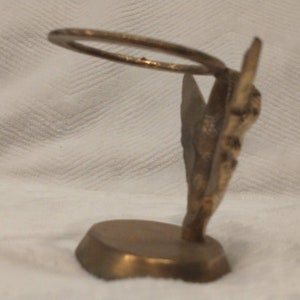 May include: A gold-toned metal decorative stand with a ring on top. The stand is shaped like a stylized butterfly with intricate details. The base is round and has a smooth surface.