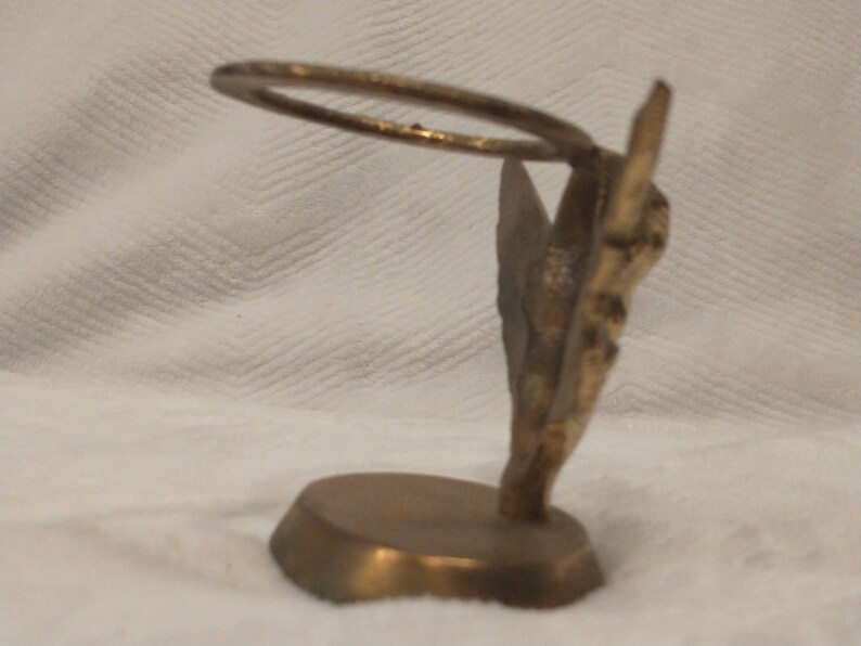 May include: A gold-toned metal decorative stand with a ring on top. The stand is shaped like a stylized butterfly with intricate details. The base is round and has a smooth surface.