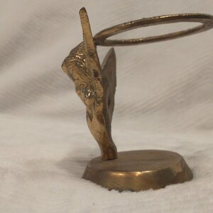 May include: A gold metal angel figurine with a halo, standing on a round base. The angel is holding a ring in its hands.