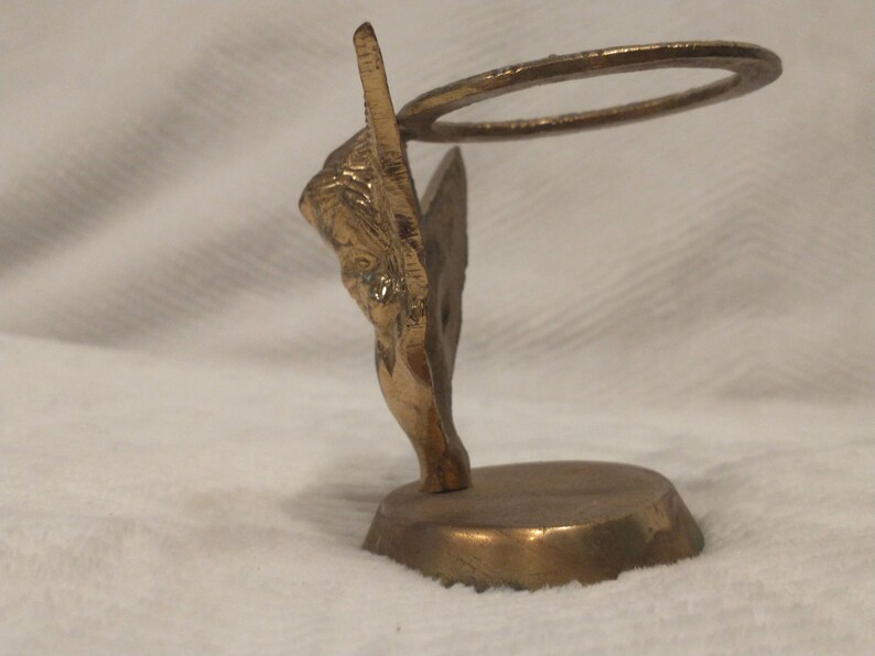 May include: A gold metal angel figurine with a halo, standing on a round base. The angel is holding a ring in its hands.