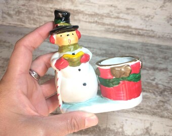May include: A ceramic Christmas snowman figurine with a black top hat, red earmuffs, and a green scarf. The snowman holds a book and is next to a red and green candle holder. The base is light blue.