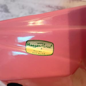May include: A close-up of a pink ceramic Haeger Floral vase. The vase has a glossy finish and a sticker with the brand name and "Made in USA". The sticker is green and gold.