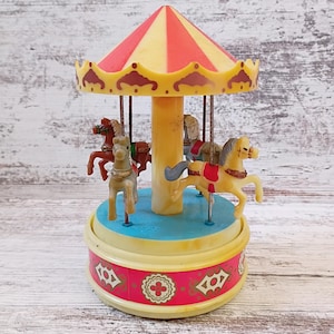 May include: A plastic carousel toy featuring a red and yellow striped canopy, a central yellow pillar, and a blue circular base. Three small molded horses in shades of brown, gray, and cream are attached to metal poles, with a pink decorative band.