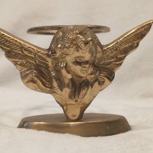 May include: A gold-toned metal angel figurine with wings outstretched, holding a round candle holder. The angel has a halo above its head and is standing on a rectangular base.