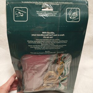 May include: A Bucilla needlecraft kit with a red canvas, gold and green yarn, and instructions. The kit is packaged in a clear plastic bag with a green and white cardboard backing. The text on the back reads "With Bucilla, your needlecraft isn't just a craft. It's an art." and "First with the Finest in Art Needlecraft... Since 1867."