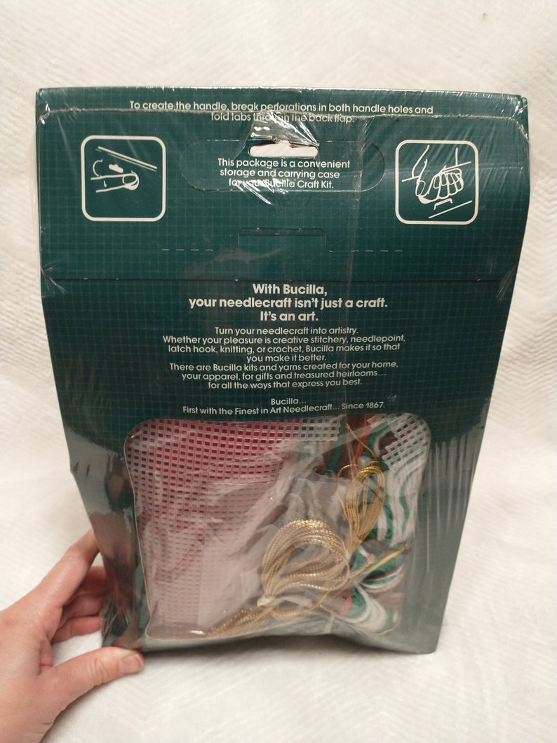May include: A Bucilla needlecraft kit with a red canvas, gold and green yarn, and instructions. The kit is packaged in a clear plastic bag with a green and white cardboard backing. The text on the back reads "With Bucilla, your needlecraft isn't just a craft. It's an art." and "First with the Finest in Art Needlecraft... Since 1867."