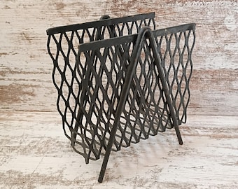 May include: A dark gray metal magazine rack with a diamond-patterned mesh design. The rack has a triangular shape with two compartments and a handle. The metal has a distressed finish, giving it a vintage look.