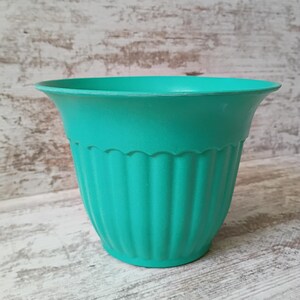 May include: A teal-colored plastic flower pot with a scalloped rim and vertical ribbed design. The pot has a flared top and a rounded base. The background is a light-colored, weathered wood surface.