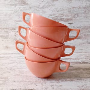 May include: A stack of five peach-colored soup bowls with handles. The bowls are stacked on top of each other, with the handles facing the same direction. The bowls have a simple, rounded design.