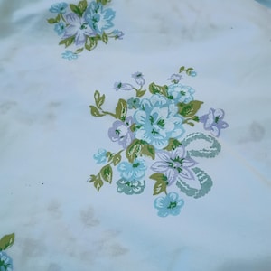 May include: A white fabric featuring a repeating floral pattern. The design consists of clusters of light blue and lavender flowers with green leaves and stems, arranged in a scattered layout across the smooth, light-colored textile surface.