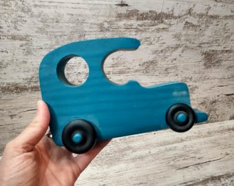 May include: A teal-colored wooden toy car with black wheels. The car has a rounded top with a circular cutout and a small, pointed front. The toy is held in a hand against a light wood-grain background.
