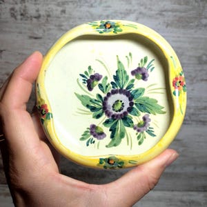 May include: A small, round, hand-painted ceramic dish with a yellow rim and a floral design. The center of the dish is off-white, featuring a bouquet of purple and green flowers. The rim is decorated with small floral accents.