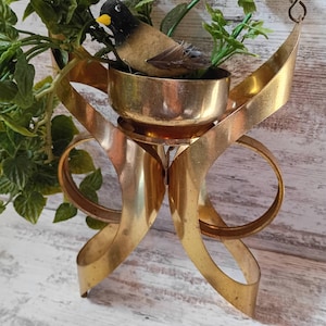 May include: A vintage brass birdcage-style decorative piece. The cage features a bowl with a small bird figurine and faux greenery. The cage has a ribbon-like design and is suspended from a hook.