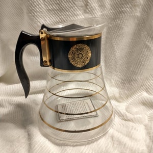 May include: A clear glass coffee pot with a black and gold band around the middle. The band has a gold design in the center. The pot has a black plastic handle and a gold lid.