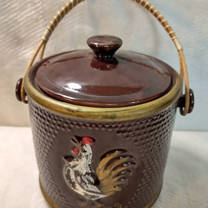 May include: Brown ceramic cookie jar with a rooster design and a woven handle. The jar has a brown lid with a gold trim and the word "Cookies" is written on the bottom.