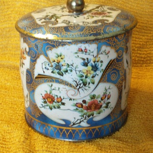 May include: A round blue and white tin with a gold trim and floral design. The lid has a gold knob.