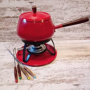 May include: A bright red fondue pot with a dark brown wooden handle and a matching lid featuring a small wooden knob. The pot sits on a black metal stand with a burner and a red base. Four fondue forks with wooden handles and colored tips are included.