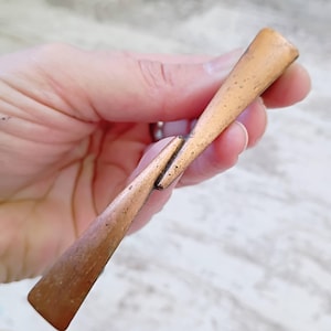 May include: A metallic, copper-toned object with a slender, elongated form that tapers toward the center. The surface features a textured, weathered appearance with visible dark spots and a matte finish, showing a distinct joint or seam in the middle.
