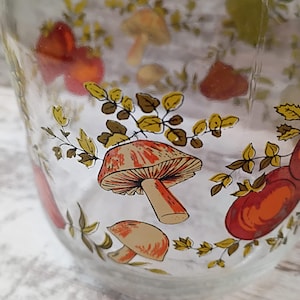 May include: Clear glass bowl with a whimsical mushroom design. The bowl features colorful illustrations of mushrooms in shades of red, orange, and tan, along with green and brown foliage. The design is repeated around the bowl's exterior.