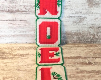 Könnte beinhalten: Handgefertigter Weihnachtsschmuck mit dem Wort "NOEL" in roten Buchstaben auf cremefarbenem Hintergrund. Die Buchstaben sind grün umrandet, mit grünen Akzenten und einer grünen Quaste unten. Oben befindet sich eine silberne Schlaufe zum Aufhängen.