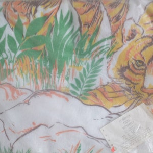 Op de afbeelding: Een witte stof met een tijgerprint in oranje, geel en bruin. Het gezicht van de tijger is zichtbaar, en de stof is omgeven door groene bladeren en gras. De stof is verpakt in een doorzichtige plastic zak met een wit etiket waarop "TIGER BUTTERFLY 72" x 150 cm staat.
