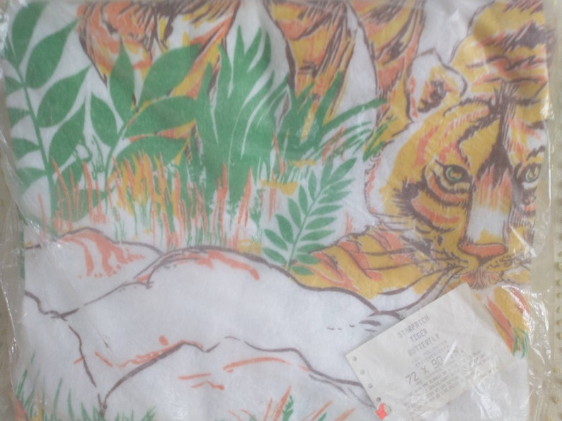 Op de afbeelding: Een witte stof met een tijgerprint in oranje, geel en bruin. Het gezicht van de tijger is zichtbaar, en de stof is omgeven door groene bladeren en gras. De stof is verpakt in een doorzichtige plastic zak met een wit etiket waarop "TIGER BUTTERFLY 72" x 150 cm staat.