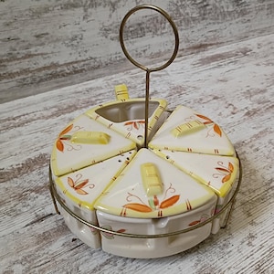 May include: A ceramic serving set consisting of five wedge-shaped containers arranged in a circle within a brass-colored metal wire frame. Each white ceramic piece features yellow edges and orange floral accents, topped with a small yellow handle.