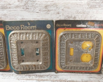 May include: Two silver-colored wall plates with decorative borders. The plates are packaged on cardboard with the text "Deco Room" printed on them. The plates are for light switches and electrical outlets.